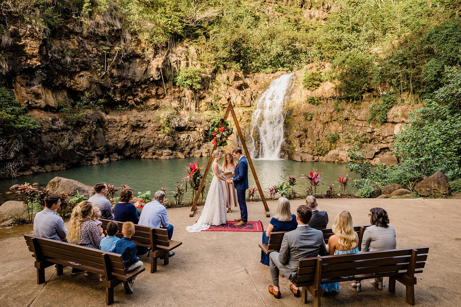 Couple stands in front of waterfall before guests during wedding ceremony.